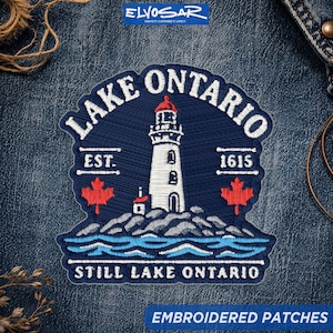 May include: A navy blue embroidered patch featuring a white lighthouse with a red top, situated on a rocky shore above blue waves. The text reads LAKE ONTARIO, EST. 1615, and STILL LAKE ONTARIO, accompanied by two red maple leaf icons on each side.