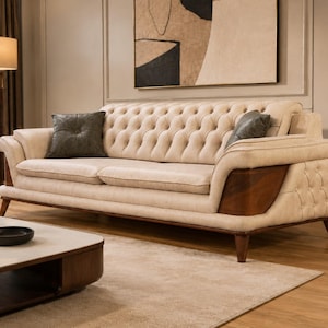 May include: A beige sofa with a tufted backrest, rolled arms, and dark wood side panels. The sofa features two seat cushions and two dark gray throw pillows. It is supported by tapered wooden legs and has a textured, light-colored fabric upholstery.