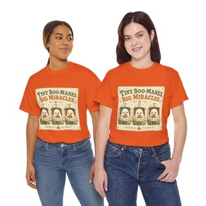May include: A bright orange short-sleeved t-shirt featuring a graphic print of three small ghost figures inside glass domes, each holding a tiny pumpkin. The text above the ghosts reads, "TINY BOO-MANES, BIG MIRACLES." The design includes decorative floral accents.