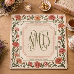Puede incluir: Un tapete de mahjong cuadrado con un centro color crema y un gran monograma verde oscuro de las letras NMB. El borde está decorado con un patrón floral de claveles rosas y hojas verdes, incluyendo instrucciones de texto para Charleston.
