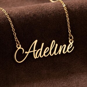 May include: A gold-toned metal necklace featuring a delicate chain and a central nameplate pendant displaying the name Adeline in a cursive script font. The pendant is attached to the chain at both ends, creating a continuous, elegant design.