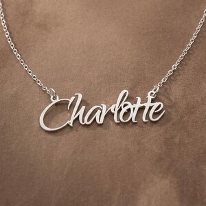 May include: A silver-toned name necklace featuring the name Charlotte written in a cursive script font. The pendant is attached to a delicate, thin metal chain that extends upwards from both sides of the nameplate, creating a classic look.