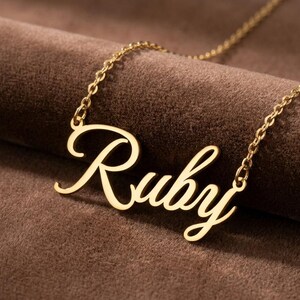 May include: A gold-toned metal necklace featuring a nameplate pendant with the name Ruby written in a cursive script font. The pendant is attached to a delicate link chain that extends from both sides of the nameplate.