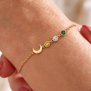 May include: A gold chain bracelet featuring a small crescent moon charm followed by three round bezel-set gemstones in yellow, clear, and green colours. The delicate metal chain connects to the moon and the stones, creating a simple link design.