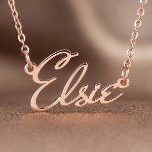 May include: A rose gold-coloured metal necklace featuring a nameplate pendant with the name Elsie written in a cursive script font. The pendant is attached to a delicate cable chain that connects to both sides of the nameplate for a balanced look.