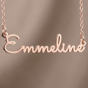May include: A rose gold-toned metal necklace featuring a nameplate pendant with the name Emmeline written in a cursive script font. The pendant is attached to a delicate link chain on both sides, creating a continuous piece of jewelry.
