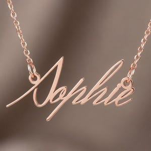 May include: A rose gold-toned metal necklace featuring a script-style nameplate pendant that spells out the name Sophie. The pendant is attached to a delicate matching chain on both sides, creating a continuous piece of jewellery.