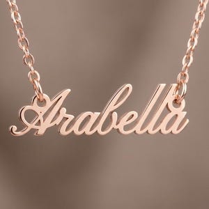 May include: A rose gold-toned necklace featuring a polished metal nameplate pendant that displays the name Arabella in a cursive script font. The pendant is suspended from a matching delicate rose gold-toned chain with circular links.