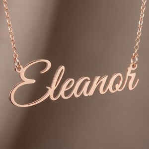 May include: A rose gold-toned name necklace featuring the name Eleanor in a cursive script font. The pendant is attached to a delicate matching chain on both sides, creating a continuous piece of jewelry with a polished metallic finish.