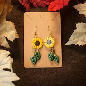 May include: A pair of dangle earrings featuring yellow sunflower charms with dark centers, attached to gold-toned metal hooks. Each earring includes two small green leaf charms suspended by short gold-toned chains below the sunflower.