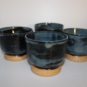May include: A set of four ceramic cups featuring a dark blue and black mottled glaze on the upper portion and a contrasting speckled beige base. Each cup has a wide, open top and a rounded body that tapers down to a solid, circular footed bottom.