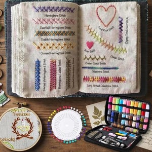 May include: An open fabric book displaying various embroidery stitches with labels including Herringbone, Deerfield, Double, Crossed, Inverted, Ladder, Coral, Zigzag, Satin, Cretan, Palestrina, and Long Armed Palestrina. Includes a hoop, pin wheel, and thread case.