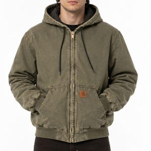 Könnte beinhalten: Eine olivgrüne Kapuzenjacke mit Reißverschluss, dunklem Innenfutter, schwarzen Kordelzügen sowie gerippten Bündchen an Ärmeln und Saum. Die Jacke verfügt über einen Frontreißverschluss und einen kleinen, quadratischen braunen Markenaufnäher.