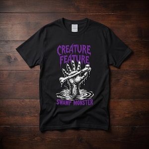 Peut inclure: Un t-shirt noir à manches courtes avec un motif représentant une main sortant de l'eau et tenant un os. Le texte "CREATURE FEATURE" est écrit en violet au-dessus et "SWAMP MONSTER" en violet en dessous. Le motif est en blanc et gris.