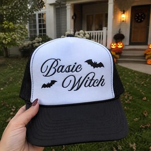 Può includere: Un cappellino trucker bianco e nero con pannello frontale bianco che riporta la scritta nera Basic Witch in carattere corsivo, decorato da due piccole sagome di pipistrelli neri. Il cappellino presenta una rete posteriore e una visiera nere.