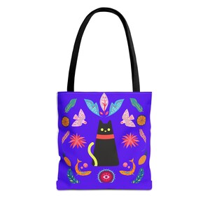 Puede incluir: Un bolso tote morado con asas negras que presenta una ilustración central de un gato negro con collar rojo y cola amarilla. El diseño incluye motivos simétricos y coloridos de pájaros, hojas, flores, peces y un símbolo de ojo central.