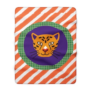 Puede incluir: Una manta rectangular con un patrón de rayas diagonales de color naranja y blanco tipo pincelada. En el centro, un gran círculo verde con patrón de cuadrícula enmarca una cara de leopardo naranja estilizada con manchas negras.