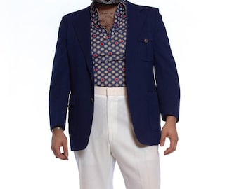 May include: A navy blue blazer with a white pocket flap and a white button. The blazer is worn over a patterned shirt with red and white circles. The person is wearing white trousers and white shoes.