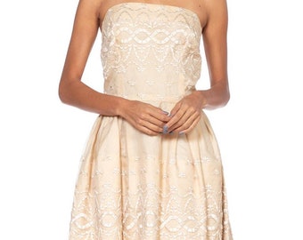 May include: A strapless, cream-coloured dress with white embroidery. The dress has a fitted bodice and a full skirt.