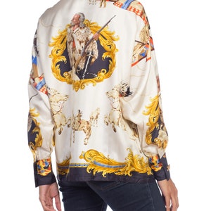 May include: A white silk blouse with a gold and blue print of a Native American man on horseback. The print also features a gold ornate frame and other Native American imagery.