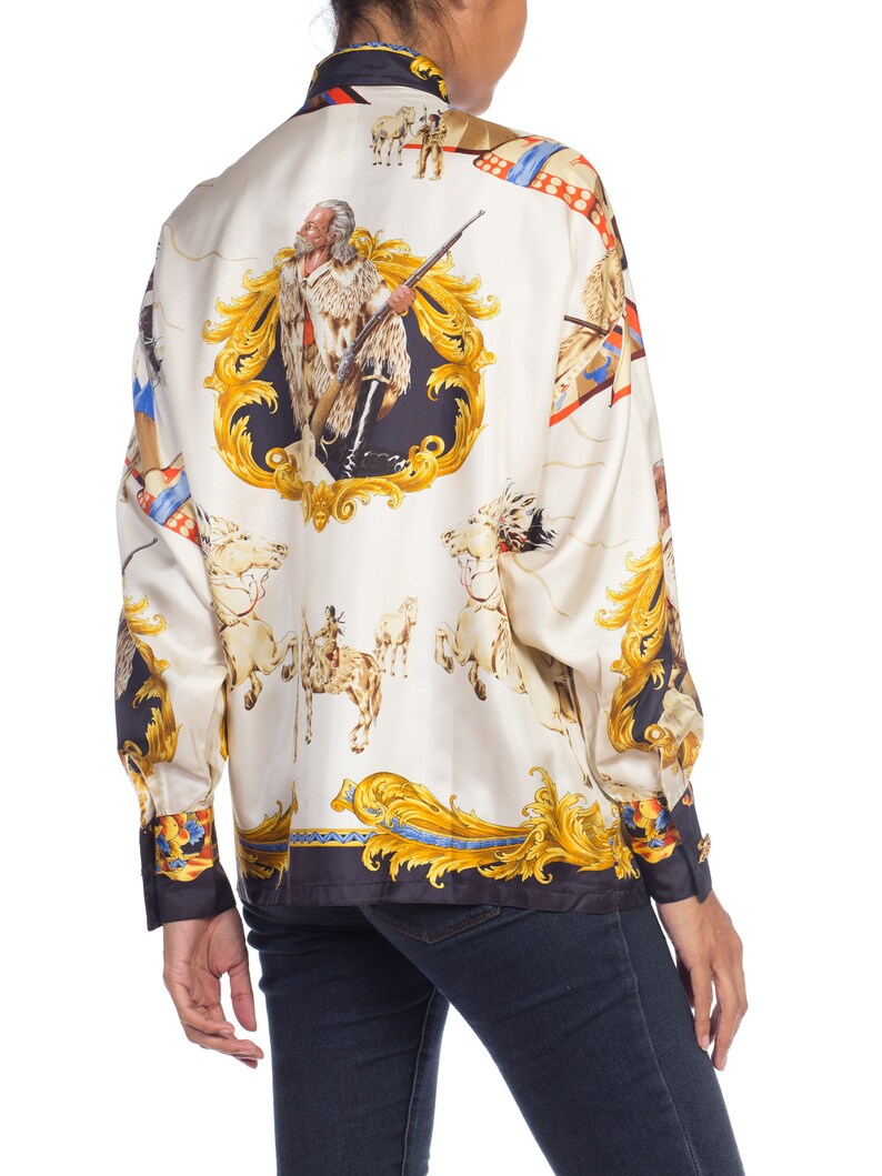 May include: A white silk blouse with a gold and blue print of a Native American man on horseback. The print also features a gold ornate frame and other Native American imagery.
