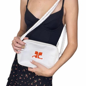 May include: White canvas crossbody bag with a rope strap and a red logo that says "AC".