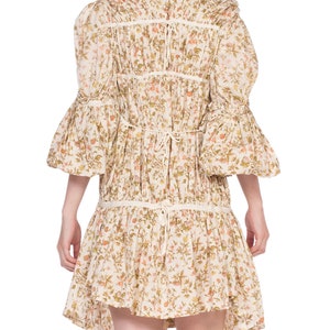 May include: A white cotton dress with a floral print and a gathered skirt. The dress has a round neckline and 3/4 sleeves with a ruffled trim. The dress is tied at the back with a ribbon.