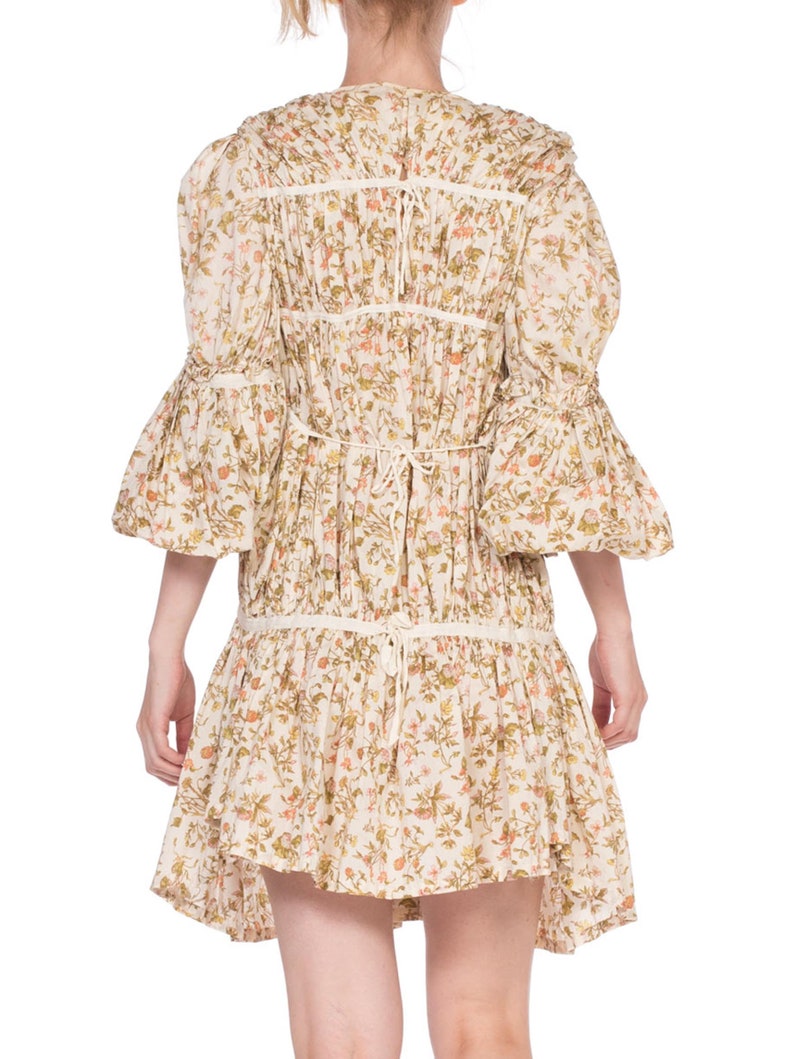 May include: A white cotton dress with a floral print and a gathered skirt. The dress has a round neckline and 3/4 sleeves with a ruffled trim. The dress is tied at the back with a ribbon.