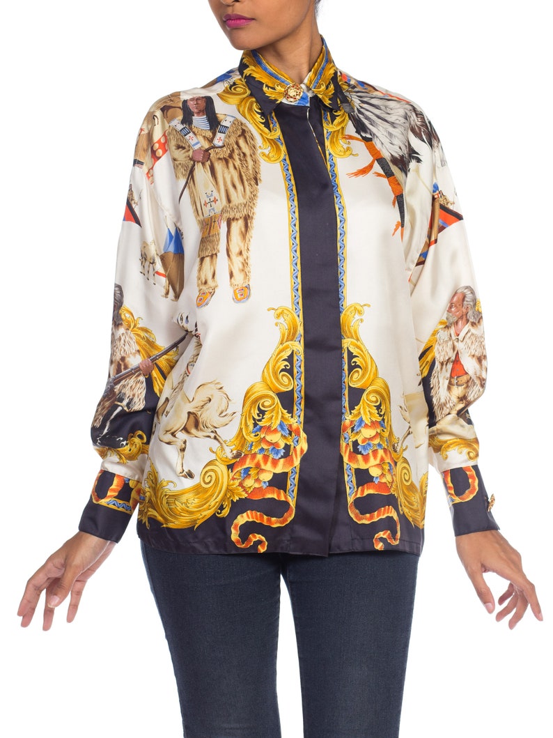 May include: A white silk button-down shirt with a black and gold print featuring Native American figures, horses, and ornate designs.