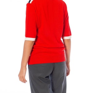 May include: A red short-sleeved sweater with white and navy blue stripes on the shoulders. The sweater is worn over gray pants.