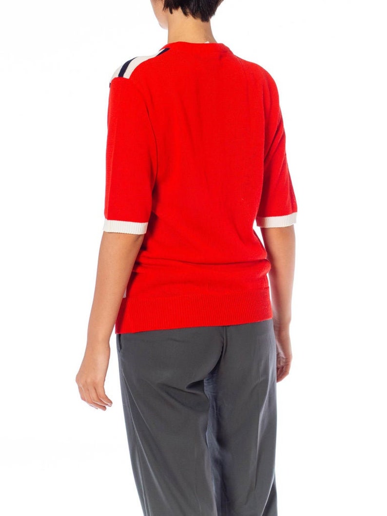 May include: A red short-sleeved sweater with white and navy blue stripes on the shoulders. The sweater is worn over gray pants.