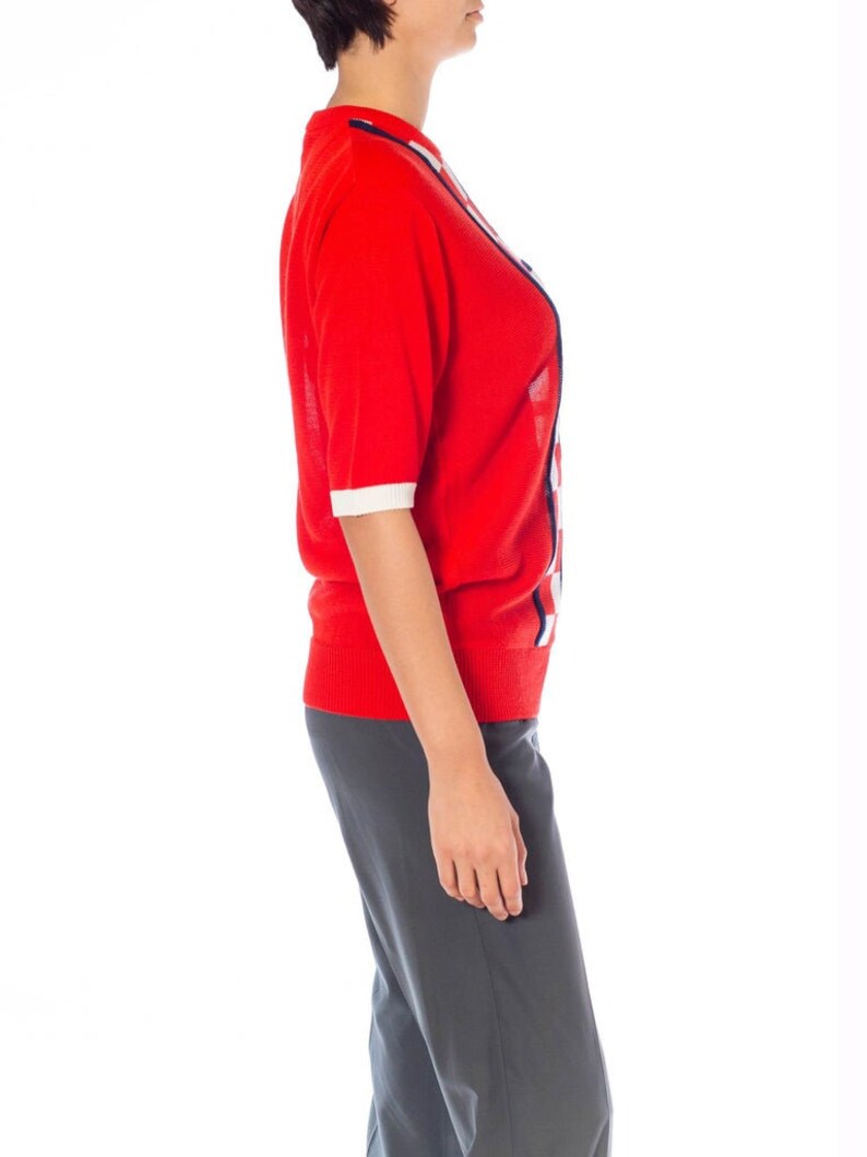 May include: A red short-sleeved sweater with white and blue geometric patterns. The sweater is made of a soft, knit fabric.