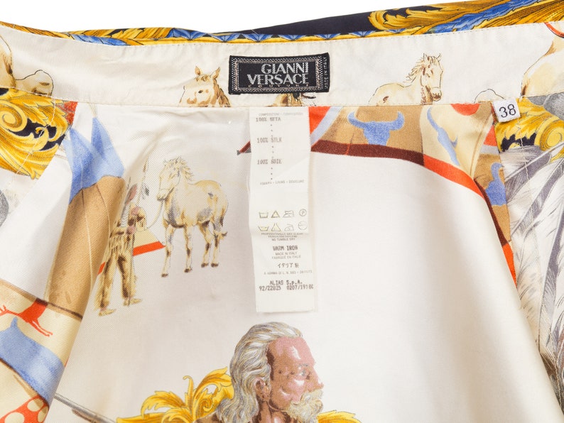 May include: A white silk shirt with a colorful print of horses, riders, and a gold design. The shirt has a label that reads "Gianni Versace" and a size tag that says "38".