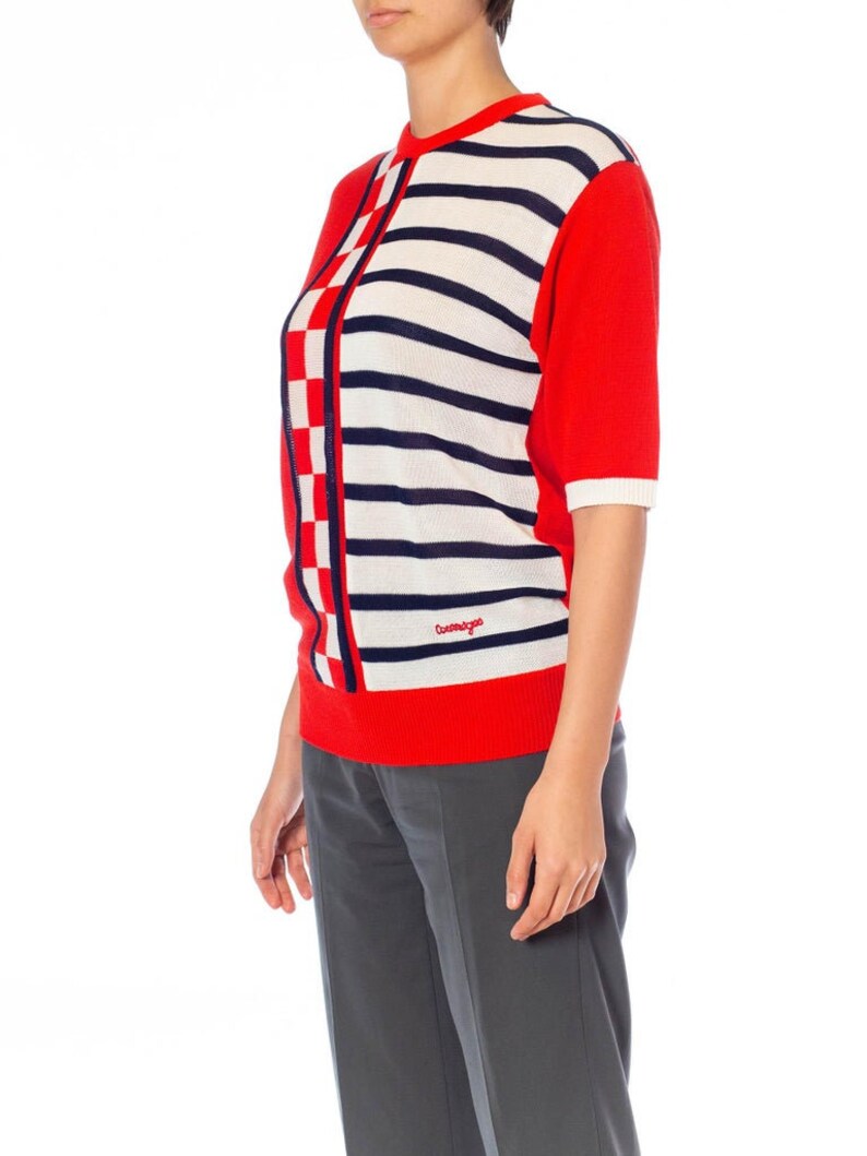 May include: A red, white, and blue striped knit sweater with a checkered pattern on the side. The sweater has short sleeves and a crew neck. The brand name "Cosmopolitan" is embroidered on the front.