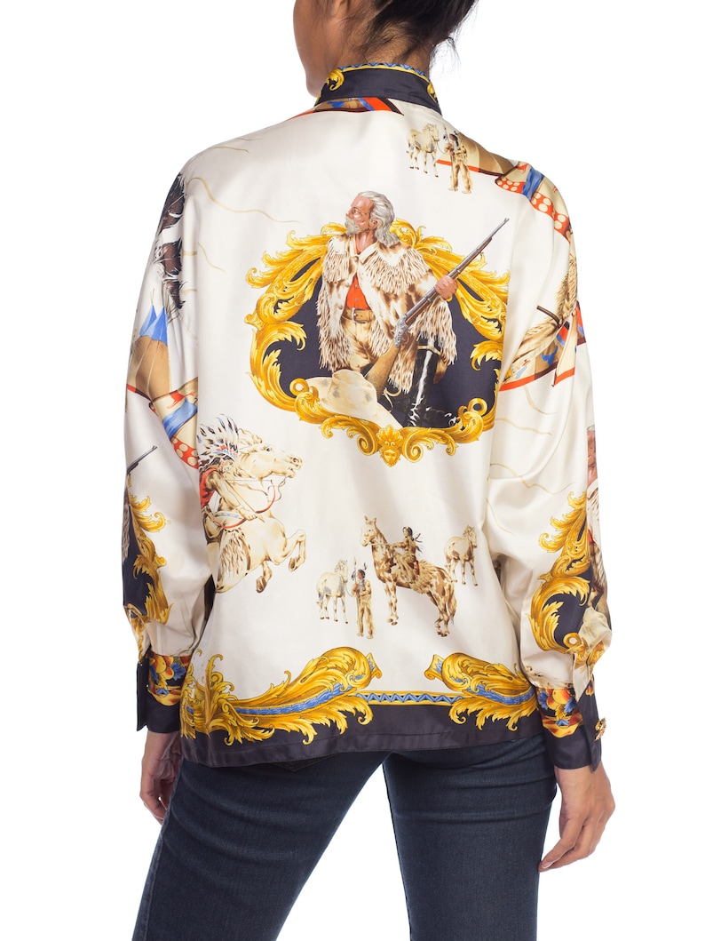 May include: A white silk blouse with a long sleeve and a button-up collar. The blouse features a colorful print of a man with a rifle, horses, and other figures. The print is in a baroque style with gold accents.