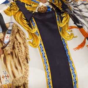 May include: A white silk shirt with a black, blue, and gold patterned design. The pattern features a Native American figure on horseback, a gold ornate design, and a blue and gold border. The shirt has a button-up collar and a gold button.
