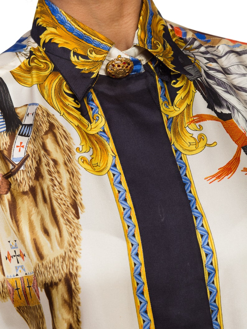 May include: A white silk shirt with a black, blue, and gold patterned design. The pattern features a Native American figure on horseback, a gold ornate design, and a blue and gold border. The shirt has a button-up collar and a gold button.