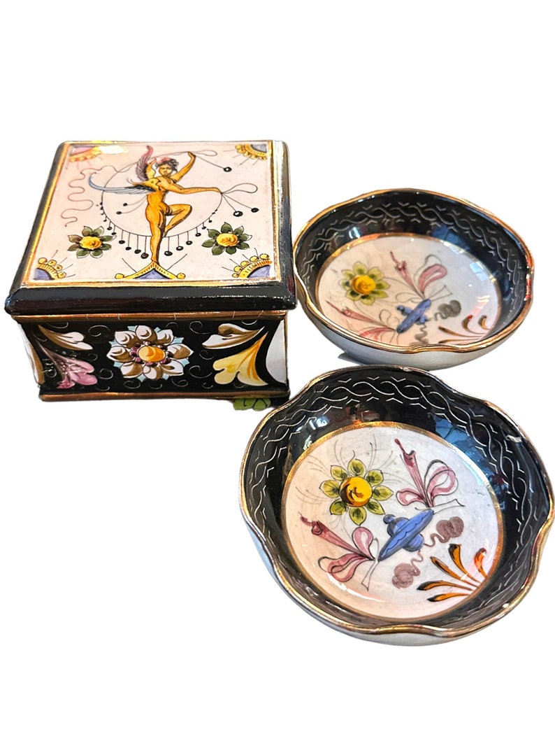 May include: A set of two ceramic bowls with black and gold trim. The bowls feature hand-painted floral designs in shades of yellow, pink, blue, and green. The larger bowl has a square box with a lid, also decorated with a hand-painted design.