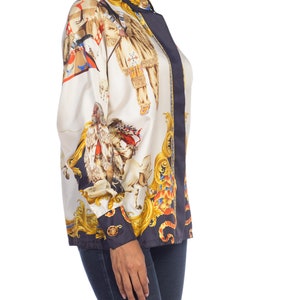 May include: A white silk blouse with a black and gold print of a scene with people and animals. The blouse has a button-up collar and long sleeves.