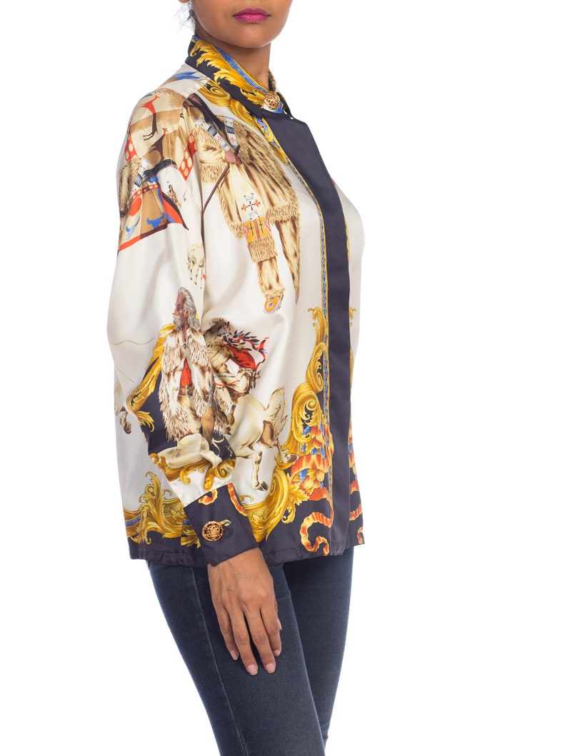 May include: A white silk blouse with a black and gold print of a scene with people and animals. The blouse has a button-up collar and long sleeves.