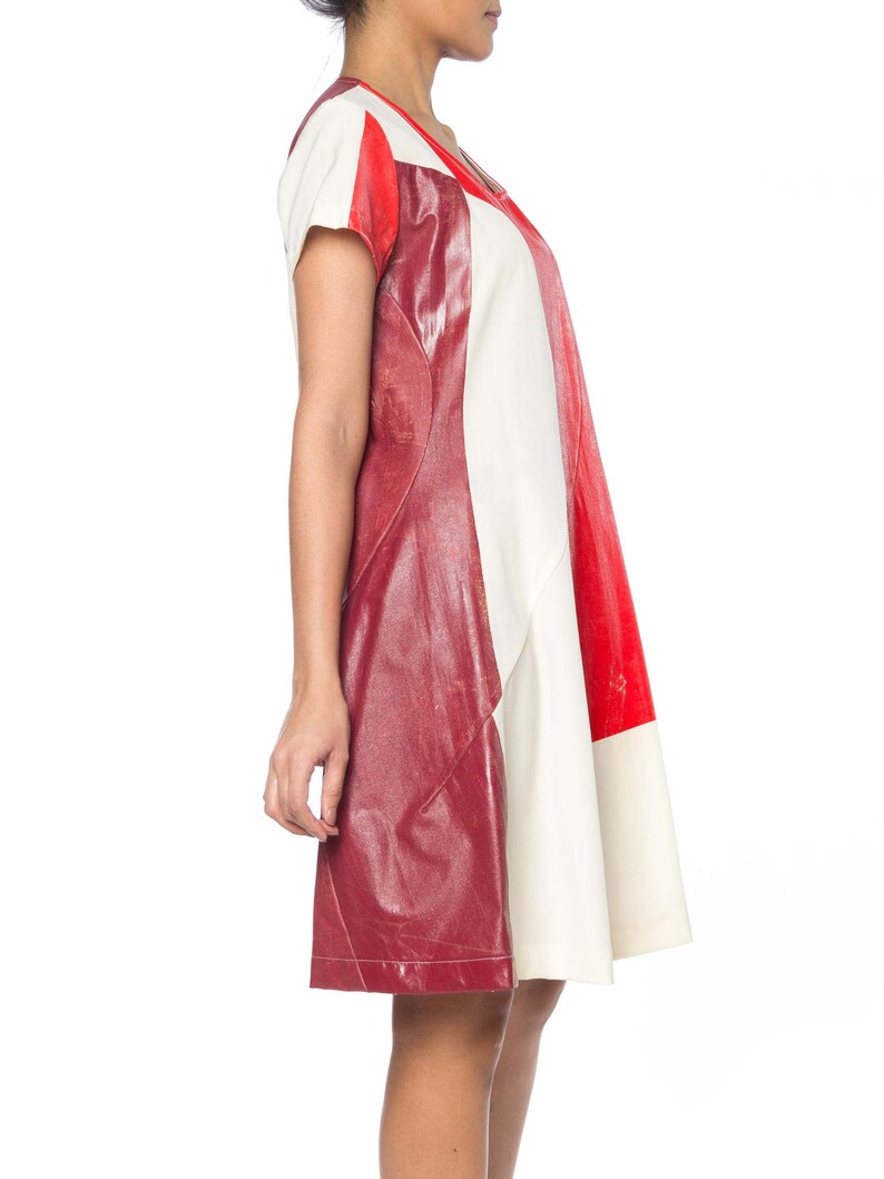 Puede incluir: Un vestido con bloques de color que presenta un dise&ntilde;o audaz con paneles rojos, blancos y burdeos. El vestido tiene mangas cortas y un escote en V. Los paneles burdeos tienen una apariencia texturizada similar al cuero, agregando un elemento &uacute;nico a la prenda.