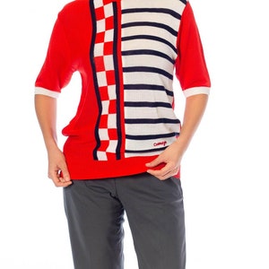 May include: Red, white, and blue short-sleeved sweater with a checkered and striped pattern. The sweater has a crew neck and the word "Comme &ccedil;a" is printed on the bottom right side.