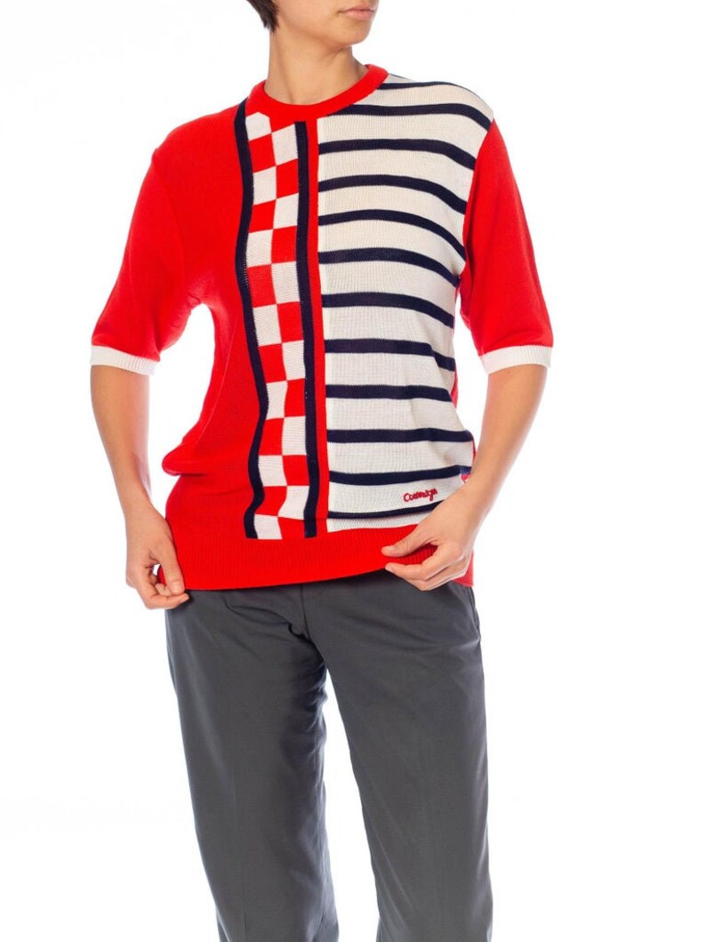 May include: Red, white, and blue short-sleeved sweater with a checkered and striped pattern. The sweater has a crew neck and the word "Comme &ccedil;a" is printed on the bottom right side.