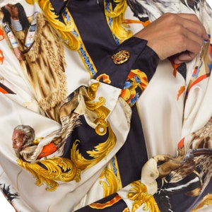 May include: A white and black silk shirt with a gold and brown print of a horse and rider. The shirt has a button-up collar and long sleeves with gold buttons.