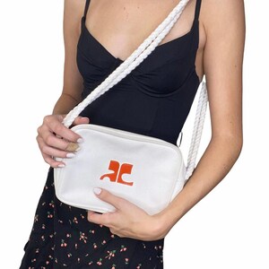 May include: White canvas crossbody bag with a rope strap and a red logo that says "AC".