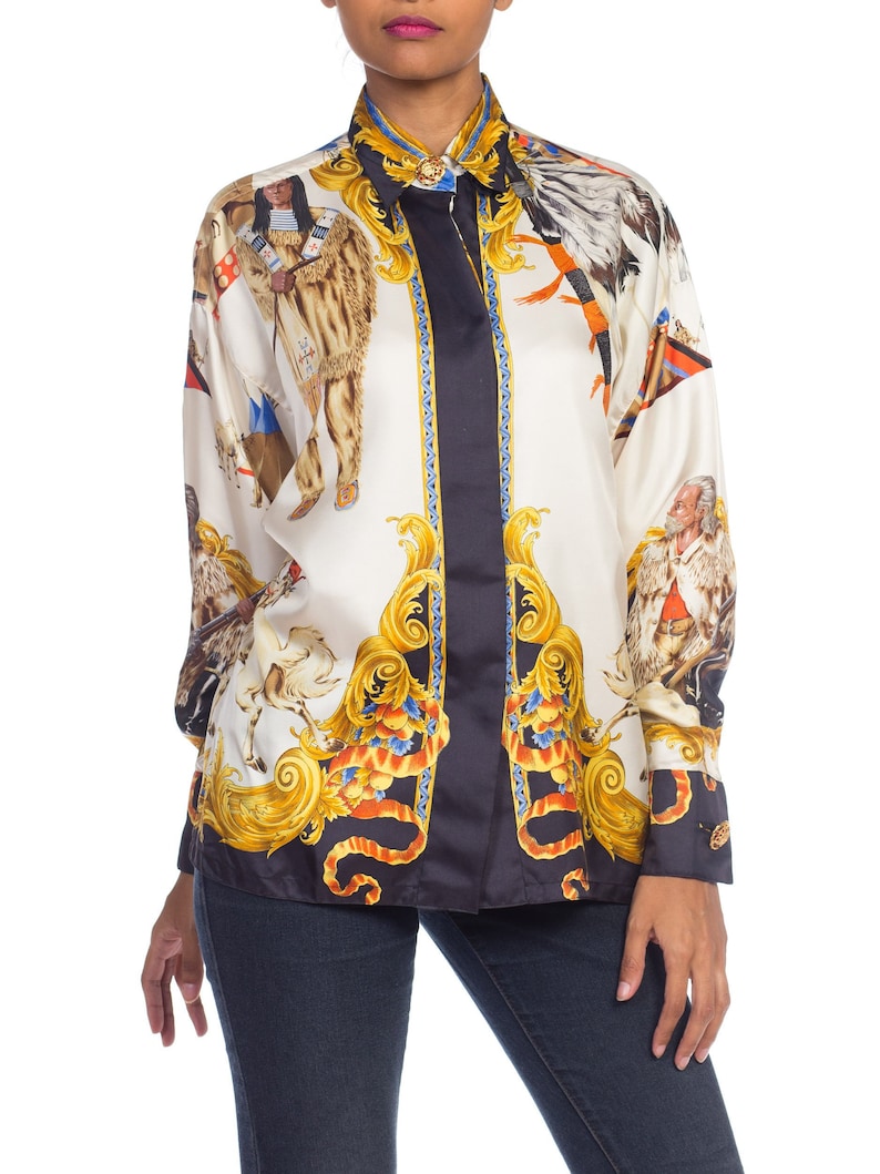 May include: A white silk button-down shirt with a black and gold print featuring Native American figures and ornate designs.