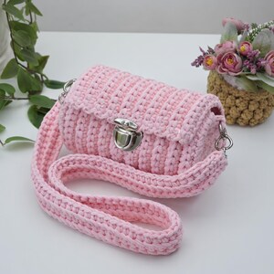 May include: A light pink crochet handbag featuring a textured ribbed pattern, a silver-toned metal push-lock closure, and a matching wide crochet shoulder strap attached with silver-toned metal swivel clasps on both sides of the bag.