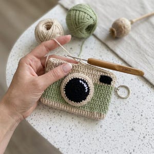 May include: A crocheted camera-shaped pouch in sage green and beige yarn with a black circular lens detail. The item features a small black rectangle accent, a metal keyring attachment, and is being worked with a 3.0mm bamboo crochet hook.