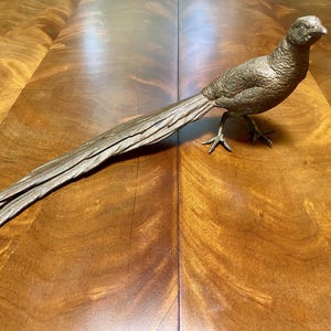 May include: A bronze-colored pheasant sculpture with a long, detailed tail. The bird is standing on three legs, with its head turned slightly to the right. The sculpture is placed on a wooden surface.