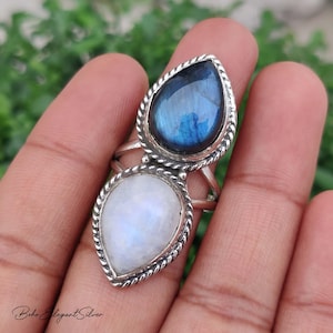 May include: A silver ring featuring two pear-shaped gemstones set in a twisted metal frame. The top stone is a dark blue labradorite with iridescent flashes, and the bottom stone is a translucent white moonstone with a soft, glowing sheen.